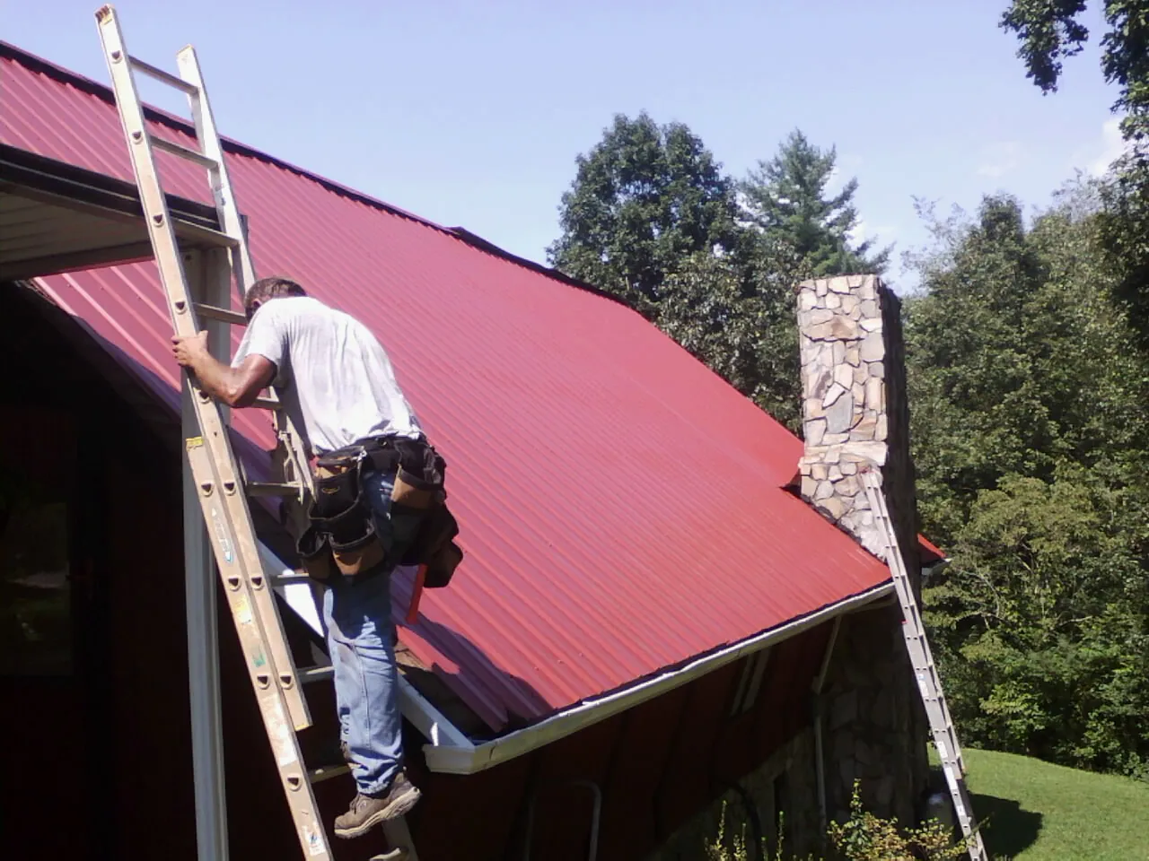 Roofer on a ladder beside a red metal roof and stone chimney — Soffit & Fascia Repair by Roofing Brothers in New Bern